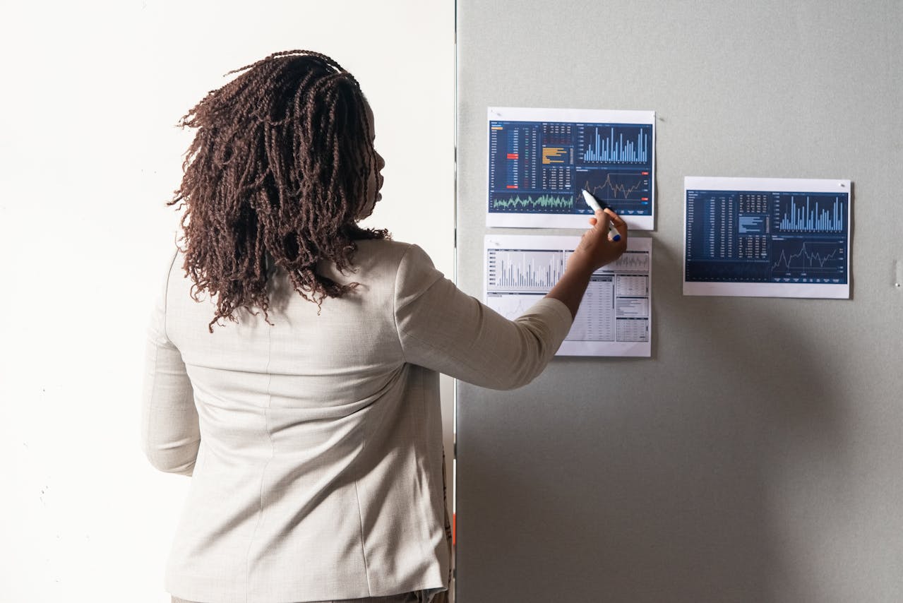 Woman with locs pointing at financial charts and data graphs pinned to a wall in an office setting.
